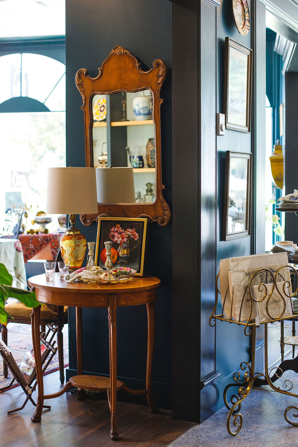 Decorative interior with a round wooden table, lamp, and framed pictures against a dark blue wall.
